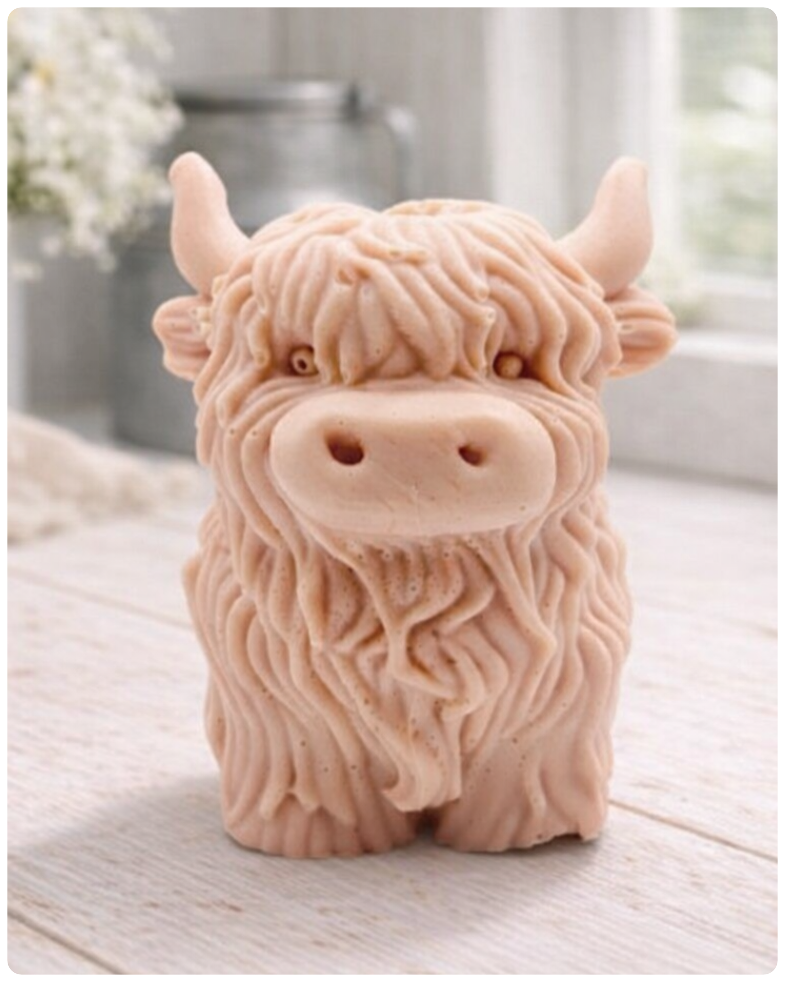 A detailed handmade vegan soap in a sculpted in the shape of a highland cow with characteristic long, wavy fur, horns, and a friendly face, displayed on a light wooden surface