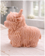 Load image into Gallery viewer, A detailed handmade vegan soap in a sculpted in the shape of a highland cow with characteristic long, wavy fur, horns, and a friendly face, displayed on a light wooden surface
