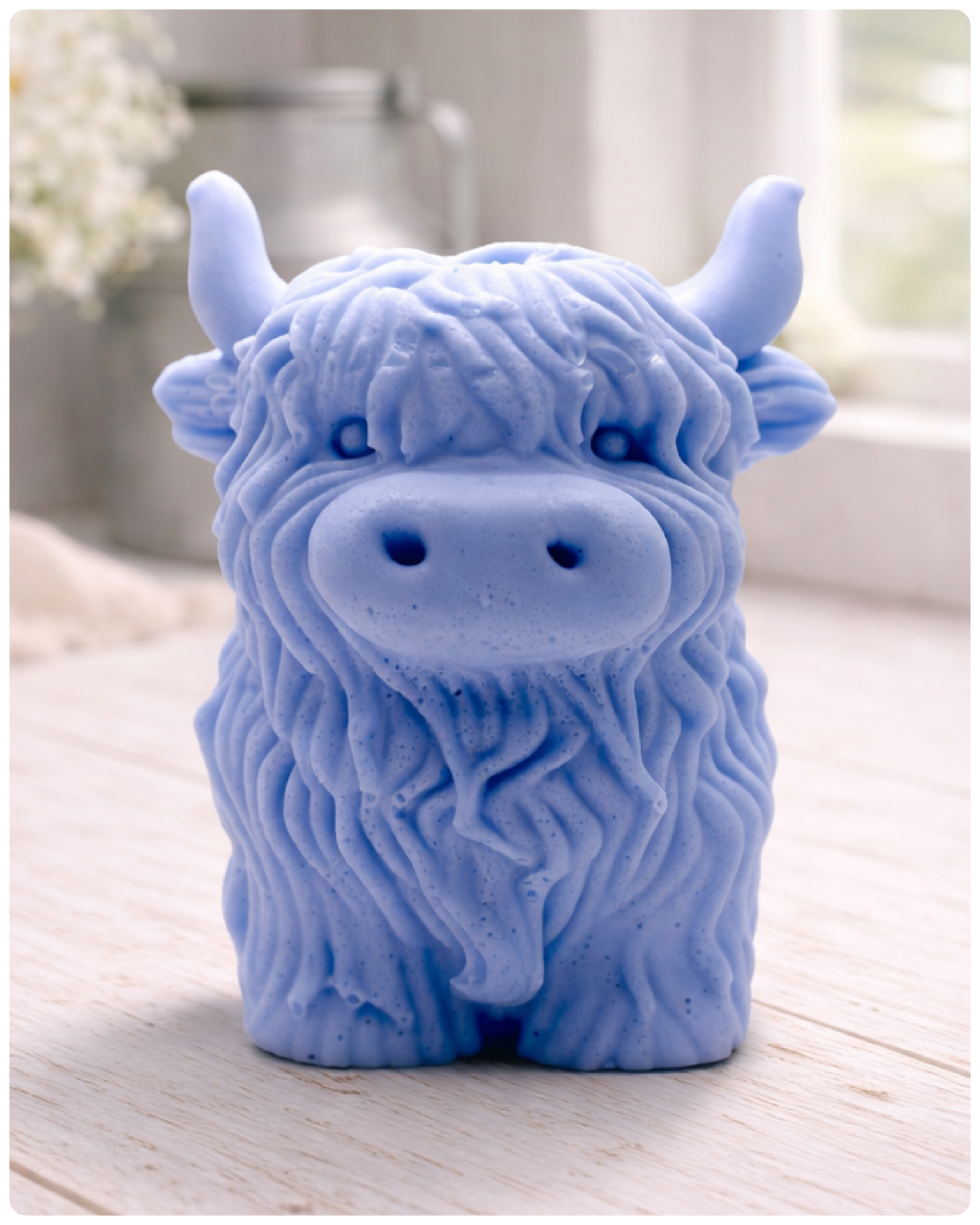 A detailed handmade vegan soap in a sculpted in the shape of a highland cow with characteristic long, wavy fur, horns, and a friendly face, displayed on a light wooden surface