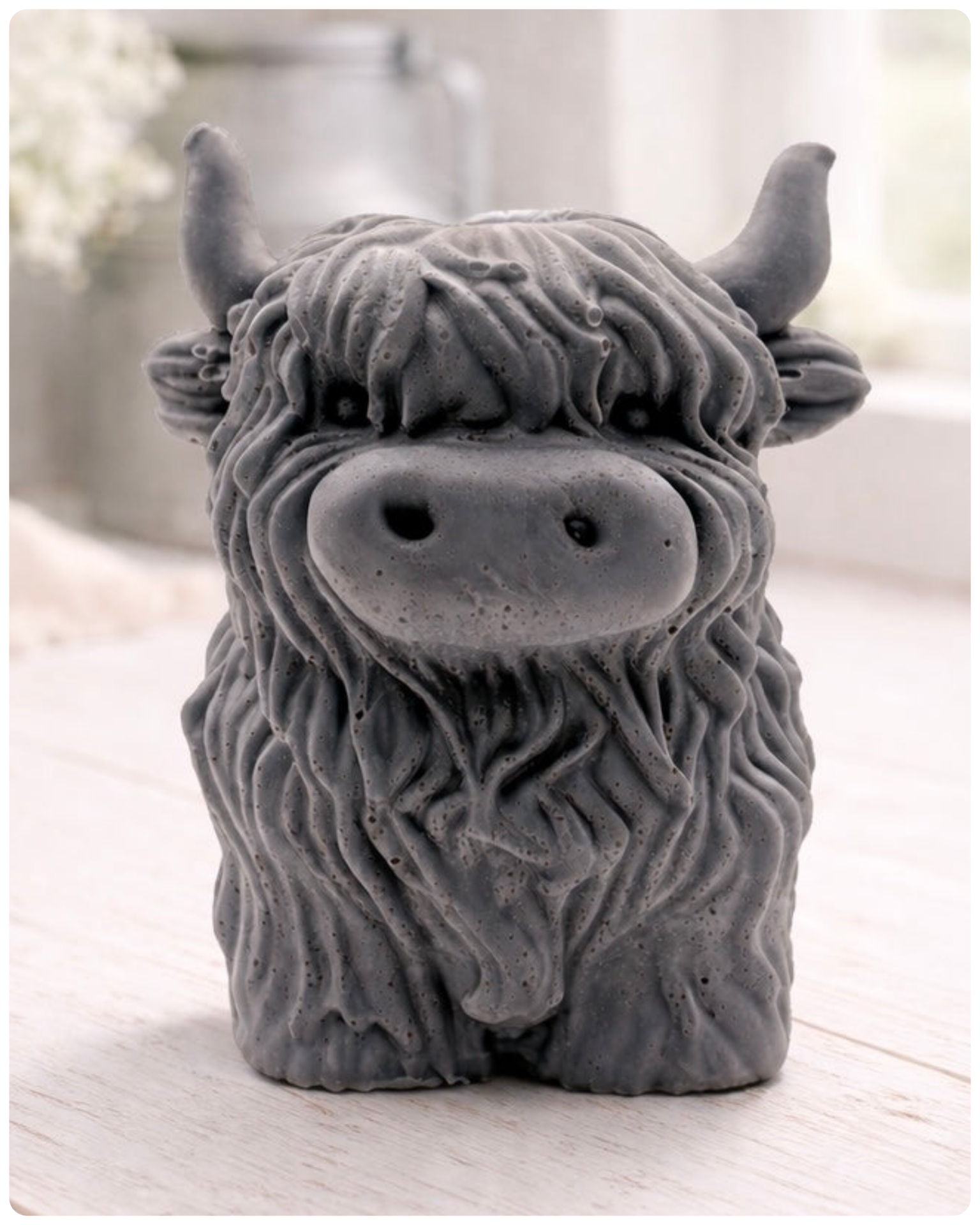 A detailed handmade vegan soap in a sculpted in the shape of a highland cow with characteristic long, wavy fur, horns, and a friendly face, displayed on a light wooden surface