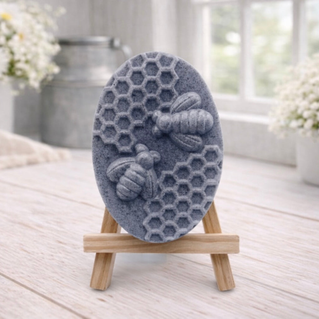handmade vegan soap featuring an oval relief sculpture of two honey bees crawling on a honeycomb pattern, displayed on a small wooden easel in a bright farmhouse decor setting.