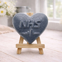 Load image into Gallery viewer, handmade vegan soap in a heart shape featuring an "NHS" relief and a heartbeat pulse line, displayed on a small wooden easel in a bright farmhouse decor setting.

