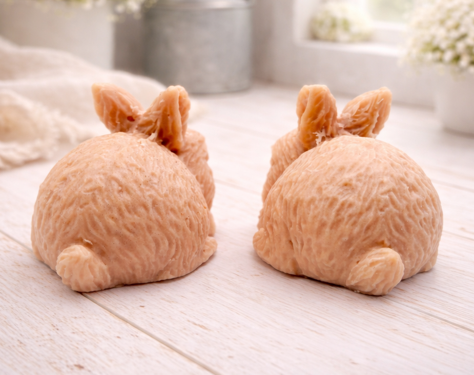 Set of two handmade vegan soaps in the shape of realistic crouching rabbits with detailed fur texture, displayed on a rustic white wooden surface in a bright farmhouse decor setting. Gift boxed. 