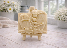 Load image into Gallery viewer, A handmade vegan soap depicting a skeleton resting between two R.I.P. gravestones, displayed on a small wooden easel.
