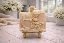 Load image into Gallery viewer, A handmade vegan soap depicting a skeleton resting between two R.I.P. gravestones, displayed on a small wooden easel.

