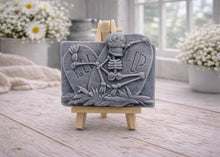 Load image into Gallery viewer, A handmade vegan soap depicting a skeleton resting between two R.I.P. gravestones, displayed on a small wooden easel.
