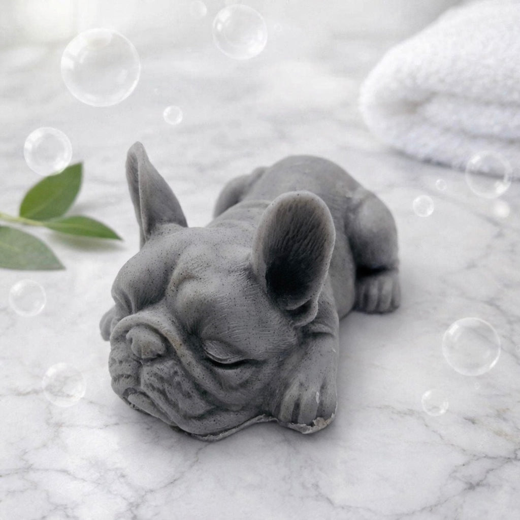 Handmade vegan soap in the shape of a realistic sleeping French Bulldog lying down, showing detailed ear texture and facial wrinkles, surrounded by bubbles in a marble bathroom setting.