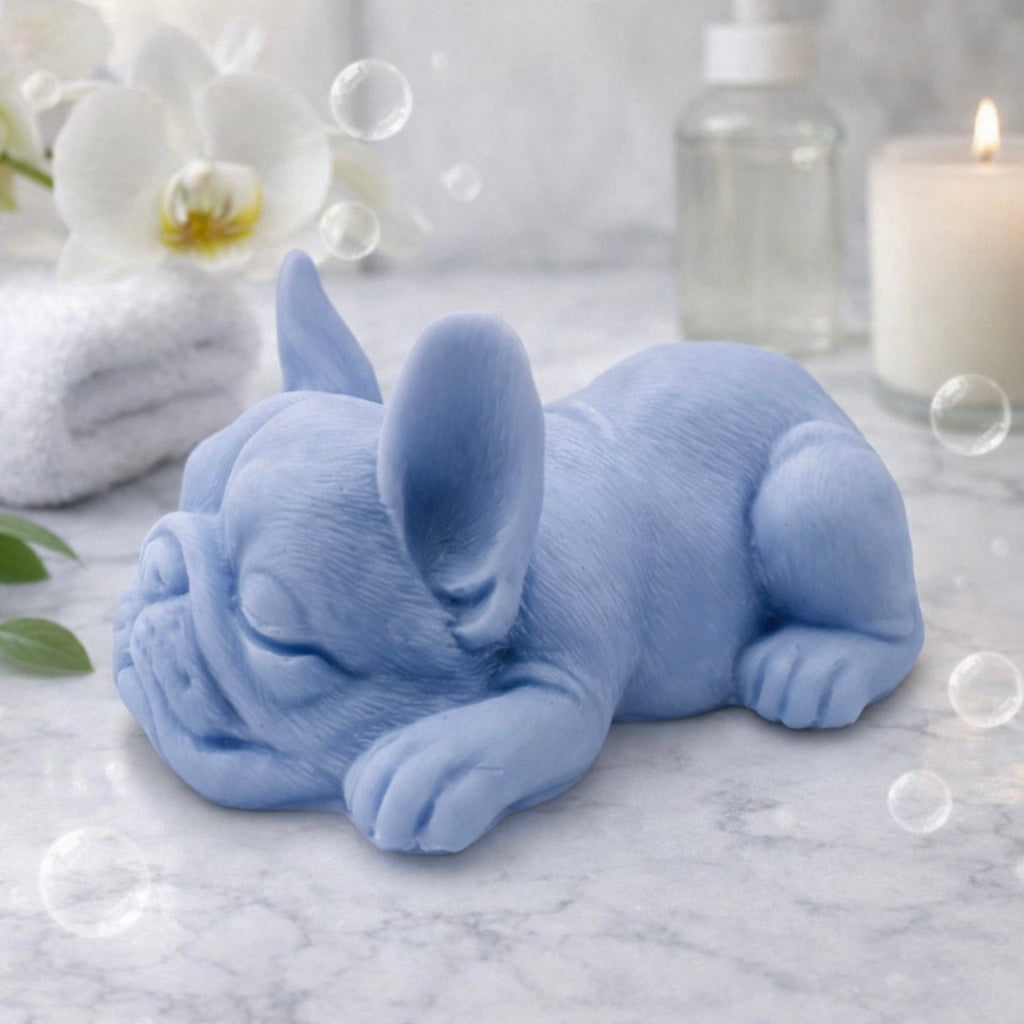 Handmade vegan soap in the shape of a realistic sleeping French Bulldog lying down, showing detailed ear texture and facial wrinkles, surrounded by bubbles in a marble bathroom setting.