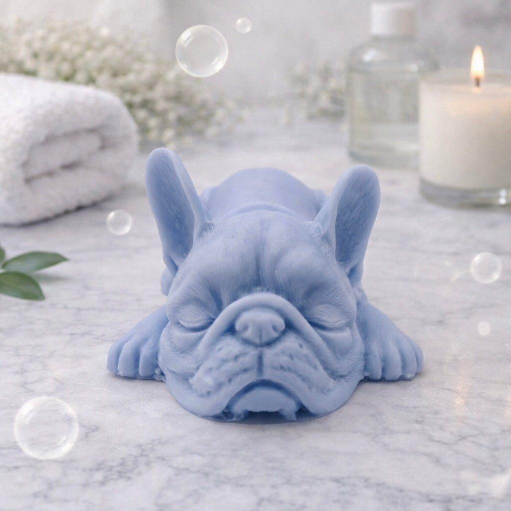 Handmade vegan soap in the shape of a realistic sleeping French Bulldog lying down, showing detailed ear texture and facial wrinkles, surrounded by bubbles in a marble bathroom setting.