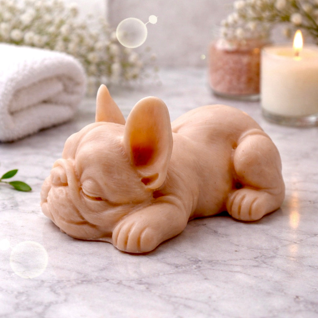 Handmade vegan soap in the shape of a realistic sleeping French Bulldog lying down, showing detailed ear texture and facial wrinkles, surrounded by bubbles in a marble bathroom setting.