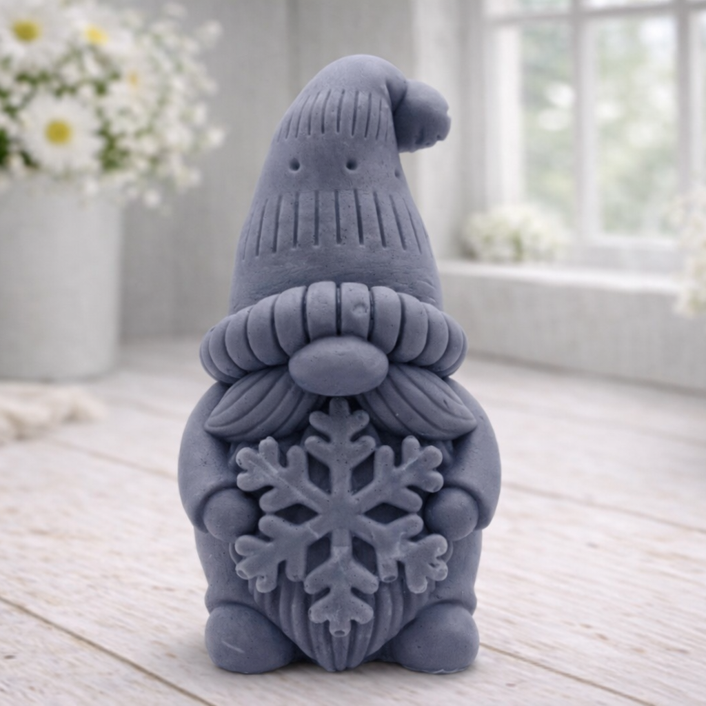 handmade vegan soap in the shape of a winter gnome sculpture holding a large snowflake, featuring a tall textured hat and a bushy moustache , displayed on a rustic white wooden surface with daisies and a milk churn in the background.