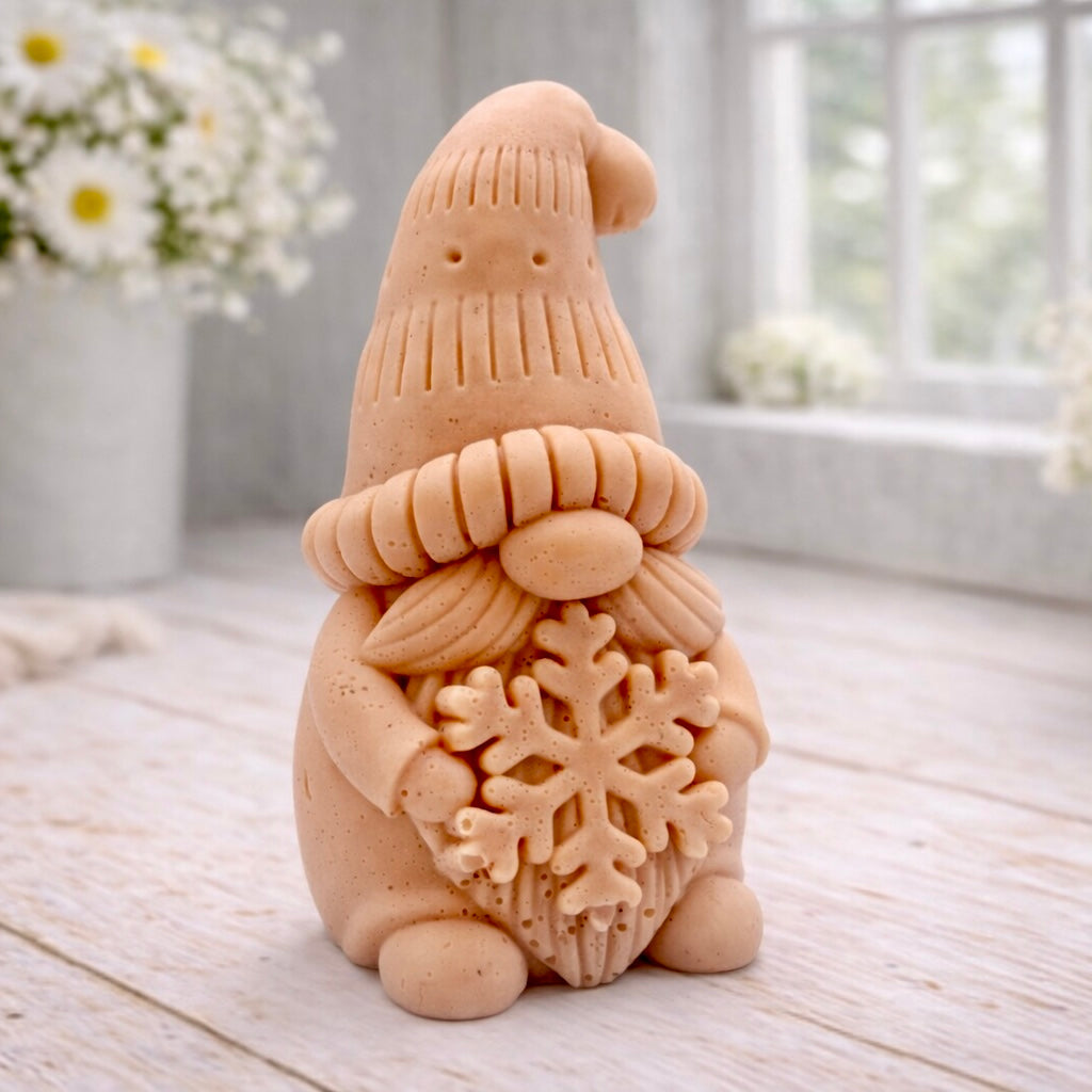 handmade vegan soap in the shape of a winter gnome sculpture holding a large snowflake, featuring a tall textured hat and a bushy moustache , displayed on a rustic white wooden surface with daisies and a milk churn in the background.