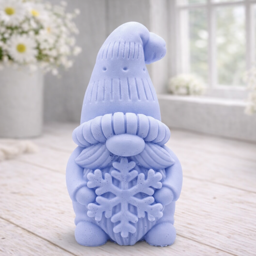handmade vegan soap in the shape of a winter gnome sculpture holding a large snowflake, featuring a tall textured hat and a bushy mustache, displayed on a rustic white wooden surface with daisies and a milk churn in the background.