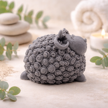 Load image into Gallery viewer, A set of three handmade vegan soaps in different sheep styles, including a dark grey smiling sheep and two lambs with detailed woolly textures, displayed on a rustic wooden surface with lavender and flowers.
