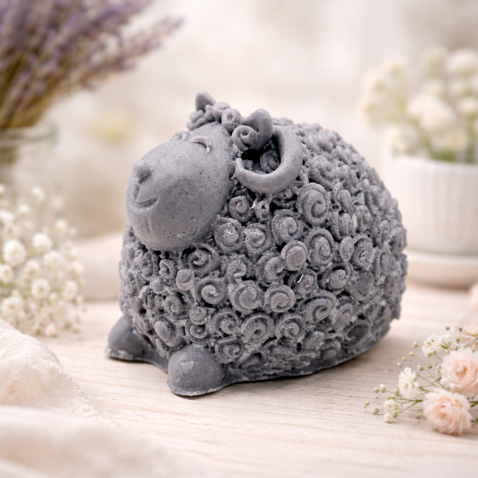A set of three handmade vegan soaps in different sheep styles, including a dark grey smiling sheep and two lambs with detailed woolly textures, displayed on a rustic wooden surface with lavender and flowers.