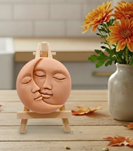 Load image into Gallery viewer, handmade vegan soap gift featuring a circular relief sculpture of a crescent moon and sun with peaceful intertwined faces, displayed on a small wooden easel in a bright farmhouse decor setting.

