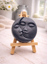 Load image into Gallery viewer, handmade vegan soap gift featuring a circular relief sculpture of a crescent moon and sun with peaceful intertwined faces, displayed on a small wooden easel in a bright farmhouse decor setting.
