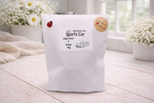 Load image into Gallery viewer, handmade vegan soap in the shape of a sleek supercar sculpture, featuring aerodynamic curves and detailed wheels, displayed on a rustic white wooden surface in a bright farmhouse decor setting.
