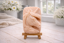 Load image into Gallery viewer, handmade vegan soap featuring a detailed unicorn head relief with a spiralled horn and flowing curly mane, displayed on a small wooden easel in a bright farmhouse decor setting.
