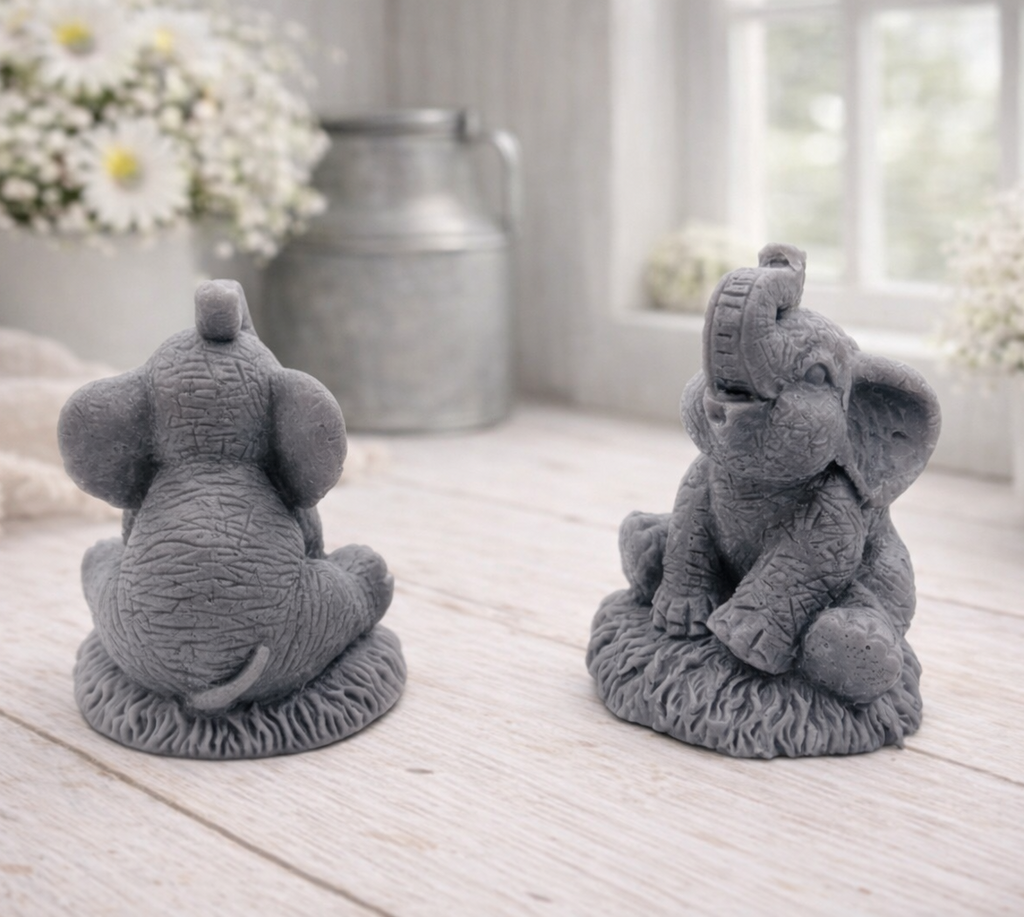 A charming gift set of two handmade vegan soaps, one sculpted as a sitting baby elephant with a trunk raised in a greeting and the other as a smaller standing calf, displayed on a light wooden surface in a bright farmhouse setting.