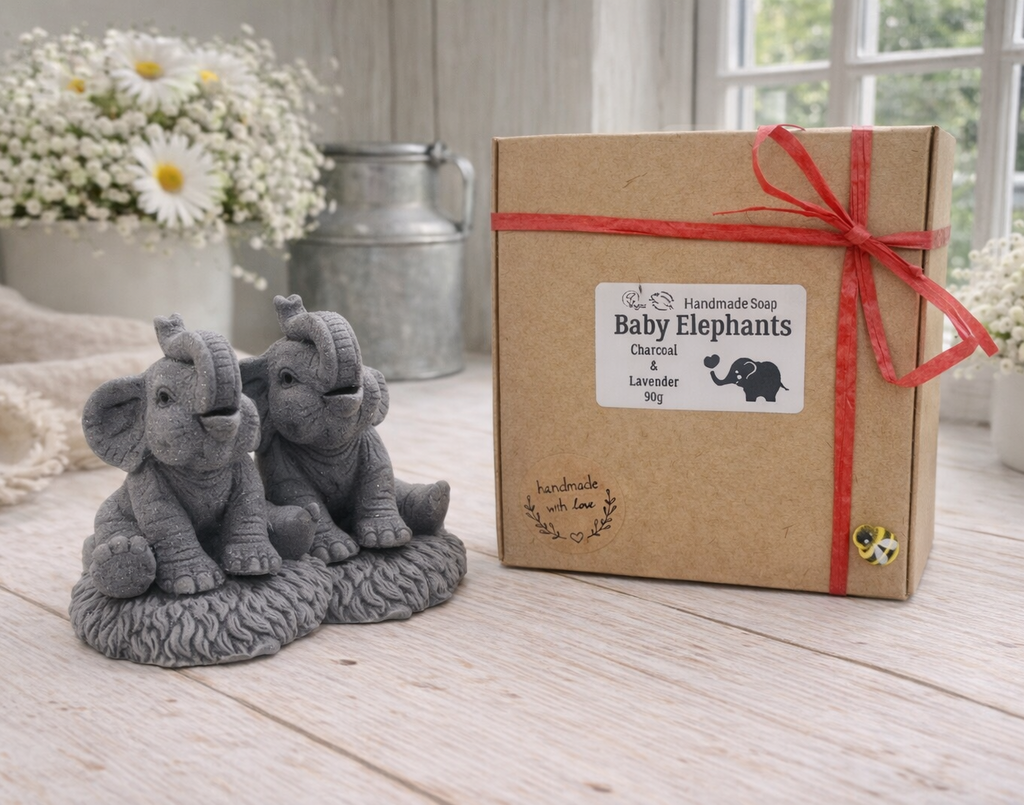 A charming gift set of two handmade vegan soaps, one sculpted as a sitting baby elephant with a trunk raised in a greeting and the other as a smaller standing calf, displayed on a light wooden surface in a bright farmhouse setting.
