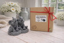Load image into Gallery viewer, A charming gift set of two handmade vegan soaps, one sculpted as a sitting baby elephant with a trunk raised in a greeting and the other as a smaller standing calf, displayed on a light wooden surface in a bright farmhouse setting.
