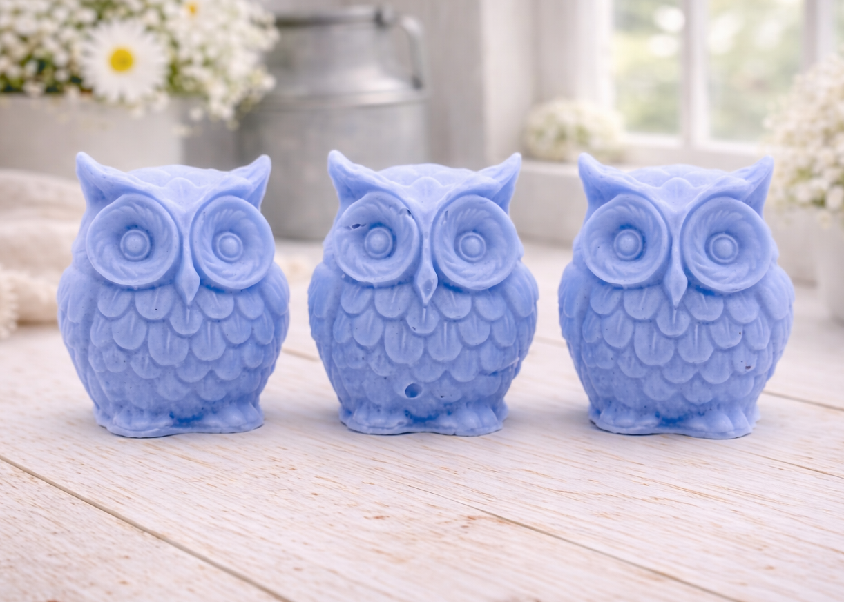 A gift set featuring three handmade vegan soaps shaped like barn owls , arranged in a row on a light wooden surface with white flowers in the background 