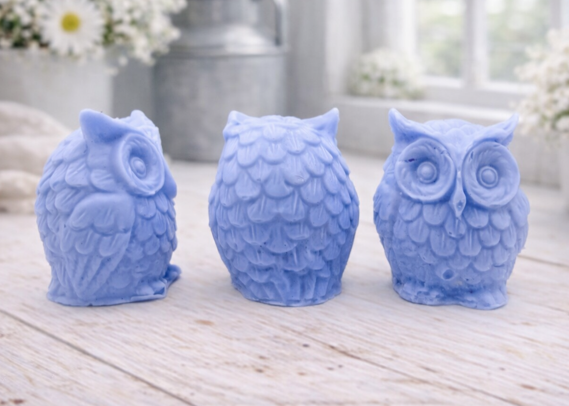 A gift set featuring three handmade vegan soaps shaped like barn owls , arranged in a row on a light wooden surface with white flowers in the background 