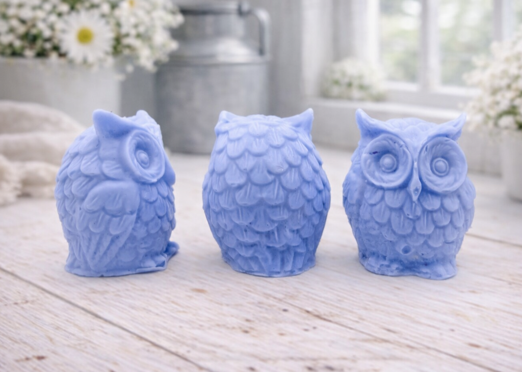 A gift set featuring three handmade vegan soaps shaped like barn owls , arranged in a row on a light wooden surface with white flowers in the background 