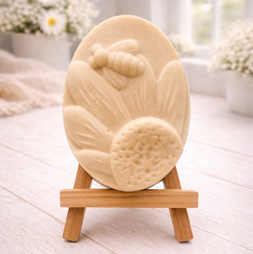 A handmade vegan soap in a natural colour, featuring an oval relief of a honeybee landing on a large sunflower with detailed petals and a textured centre, displayed on a small wooden easel.