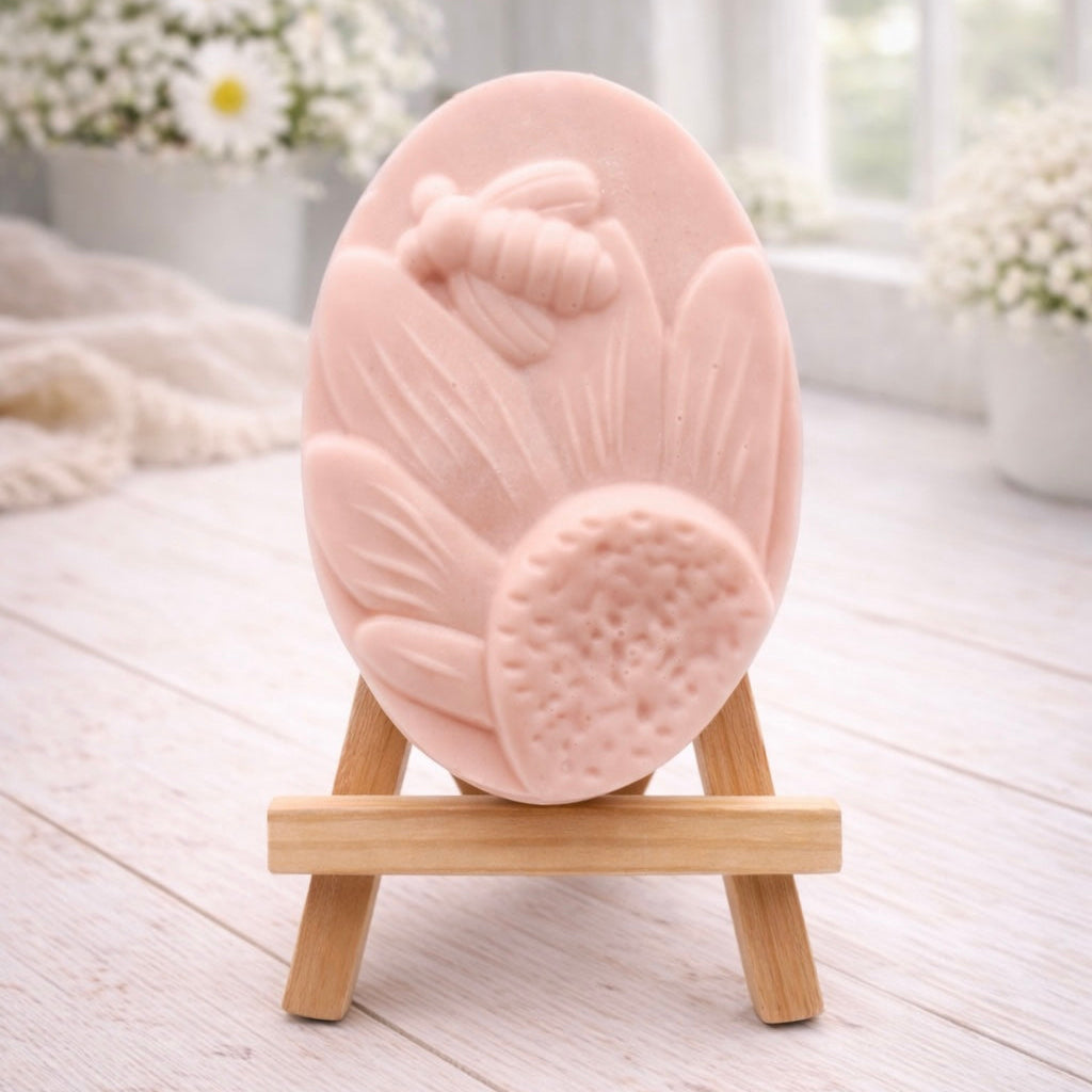 A handmade vegan soap in a natural colour, featuring an oval relief of a honeybee landing on a large sunflower with detailed petals and a textured centre, displayed on a small wooden easel.