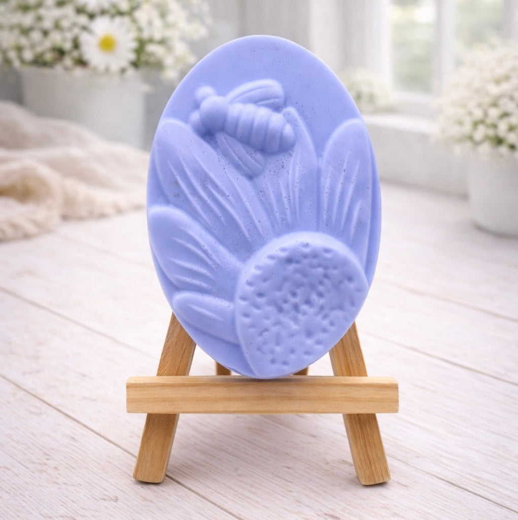 A handmade vegan soap in a natural colour, featuring an oval relief of a honeybee landing on a large sunflower with detailed petals and a textured centre, displayed on a small wooden easel.