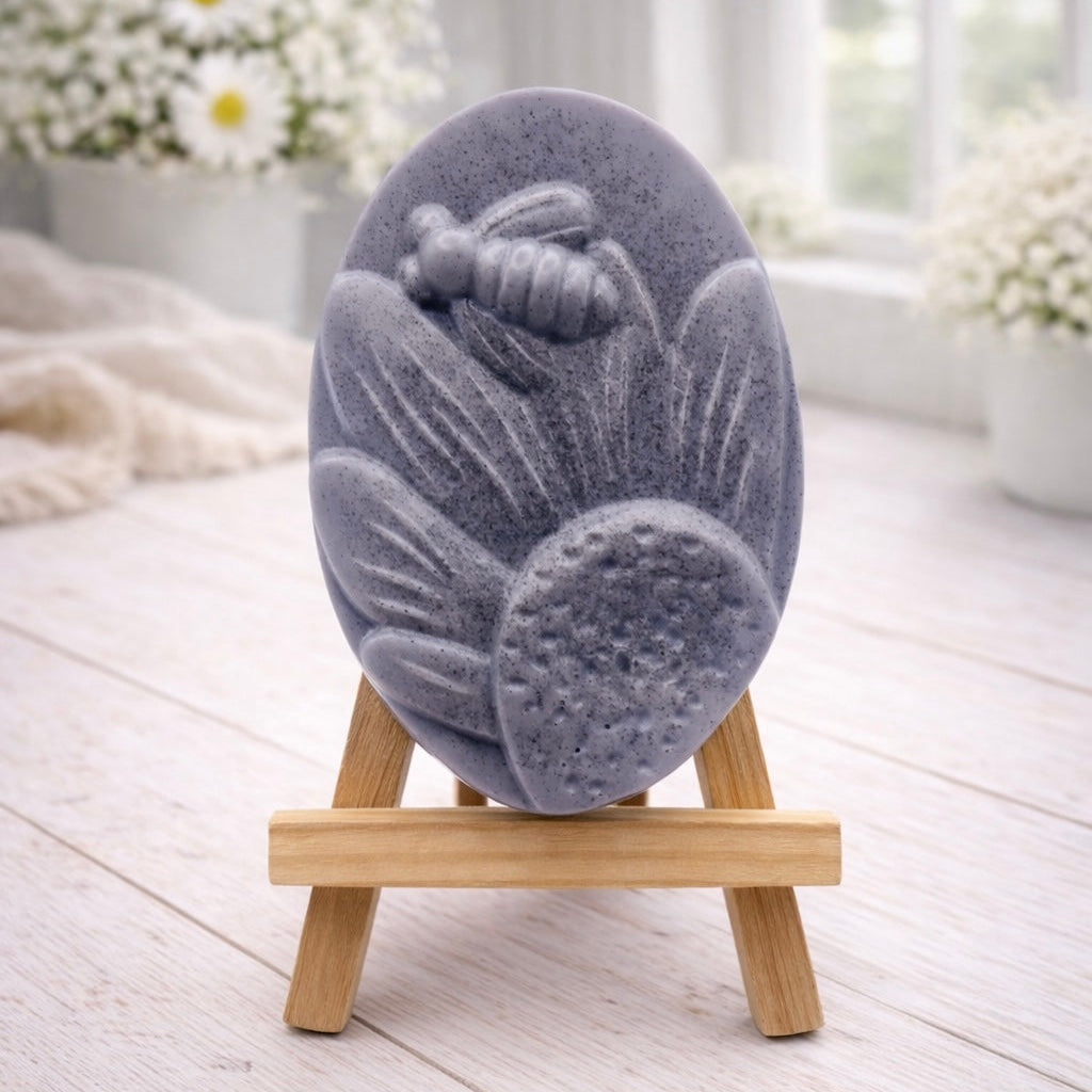 A handmade vegan soap in a natural colour, featuring an oval relief of a honeybee landing on a large sunflower with detailed petals and a textured centre, displayed on a small wooden easel.