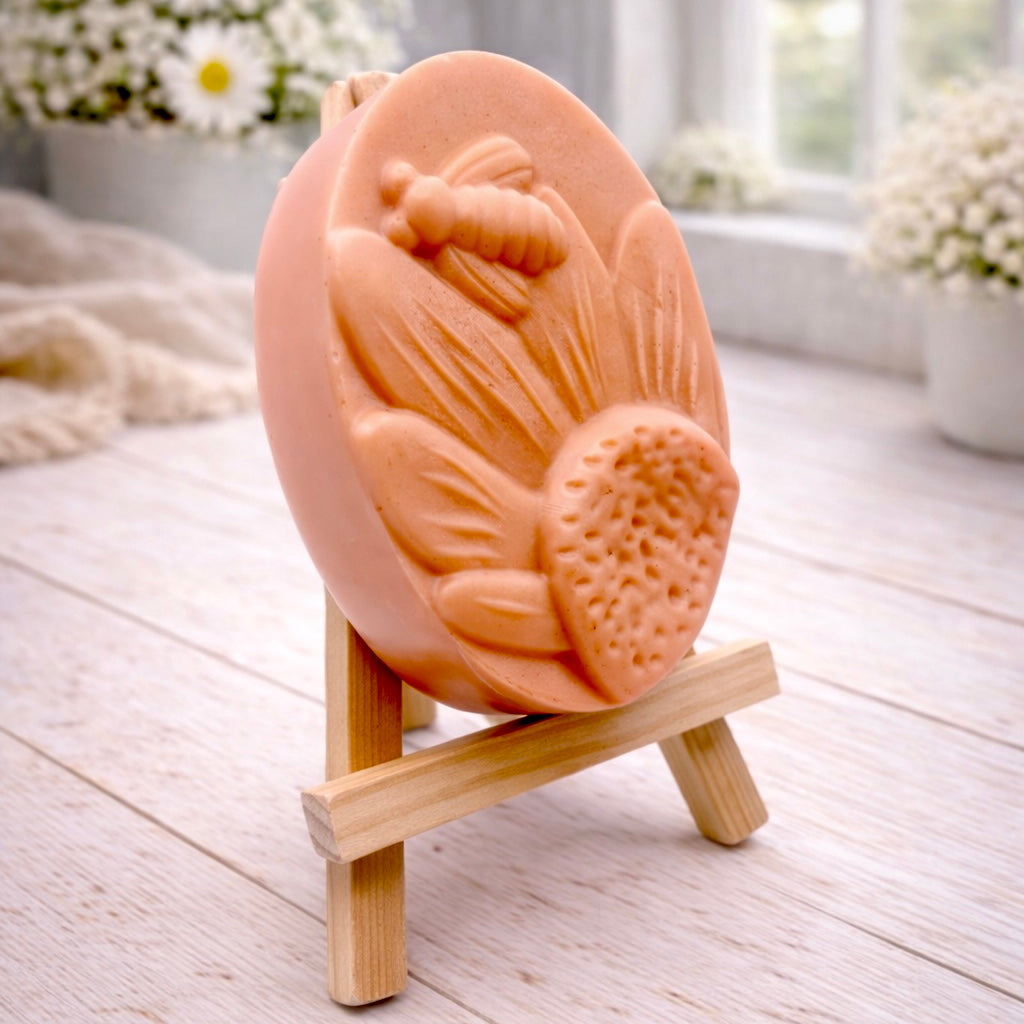 A handmade vegan soap in a natural colour, featuring an oval relief of a honeybee landing on a large sunflower with detailed petals and a textured centre, displayed on a small wooden easel.