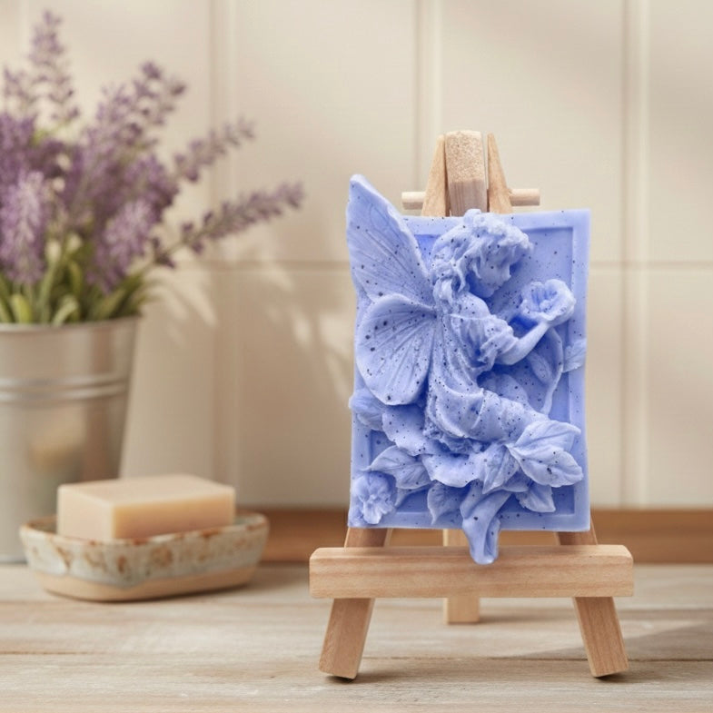 A rectangular handmade vegan soap, featuring a high-relief carving of a seated fairy with large, detailed wings holding a single blossom, displayed on a miniature wooden easel.