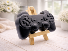 Load image into Gallery viewer, A 3D handmade vegan soap sculpted in the shape of a classic video game controller, finished in a deep charcoal black with realistic buttons and joysticks, displayed on a miniature wooden easel in a bright farmhouse setting.
