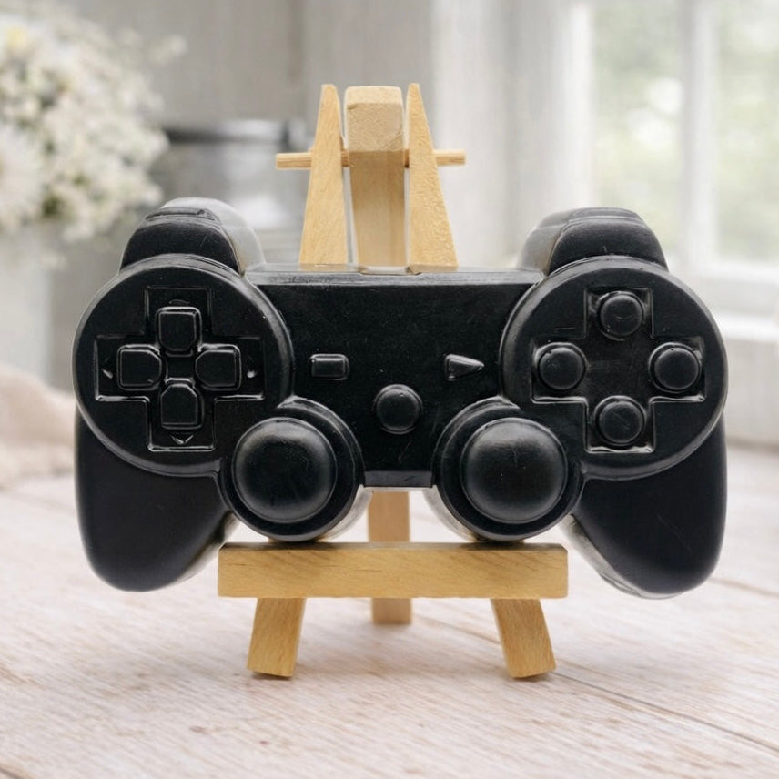 A 3D handmade vegan soap sculpted in the shape of a classic video game controller, finished in a deep charcoal black with realistic buttons and joysticks, displayed on a miniature wooden easel in a bright farmhouse setting.