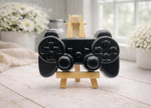 Load image into Gallery viewer, A 3D handmade vegan soap sculpted in the shape of a classic video game controller, finished in a deep charcoal black with realistic buttons and joysticks, displayed on a miniature wooden easel in a bright farmhouse setting.
