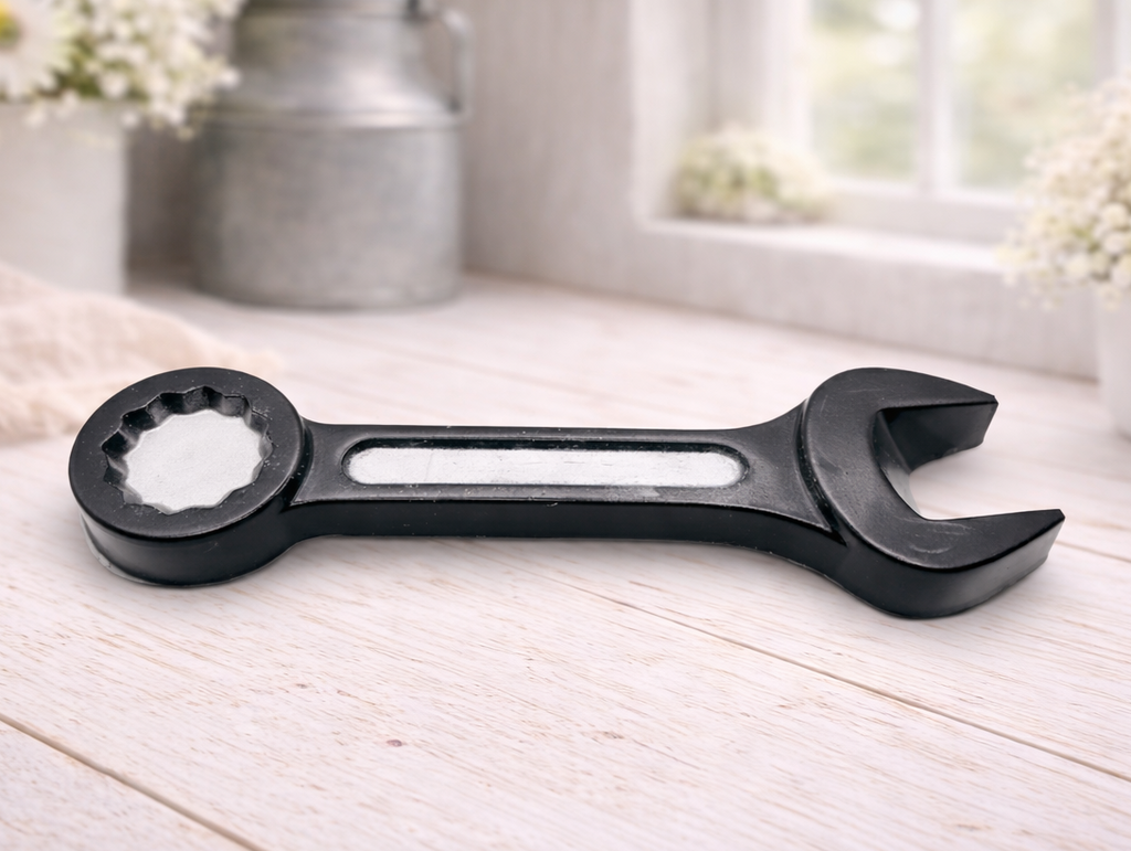 A handmade vegan soap sculpted in the shape of a realistic spanner or wrench tool, finished in a sleek charcoal black with silver-painted detailing on the handle and head, lying on a light wood surface.