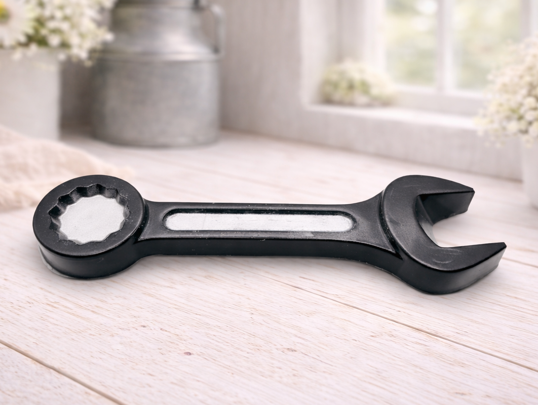 A handmade vegan soap sculpted in the shape of a realistic spanner or wrench tool, finished in a sleek charcoal black with silver-painted detailing on the handle and head, lying on a light wood surface.