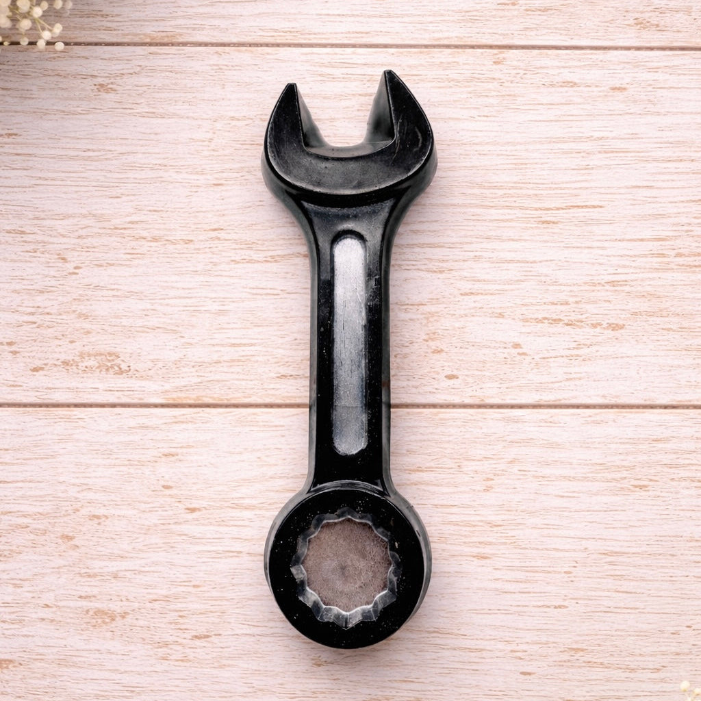 A 3D handmade vegan soap sculpted in the shape of a realistic spanner or wrench tool, finished in a sleek charcoal black with silver-painted detailing on the handle and head, lying on a light wood surface.