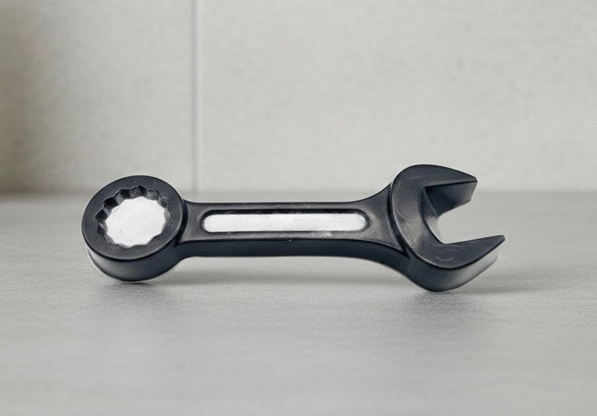 A 3D handmade vegan soap sculpted in the shape of a realistic spanner or wrench tool, finished in a sleek charcoal black with silver-painted detailing on the handle and head, lying on a light wood surface.