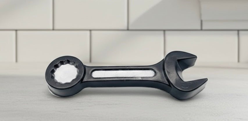 A 3D handmade vegan soap sculpted in the shape of a realistic spanner or wrench tool, finished in a sleek charcoal black with silver-painted detailing on the handle and head, lying on a light wood surface.