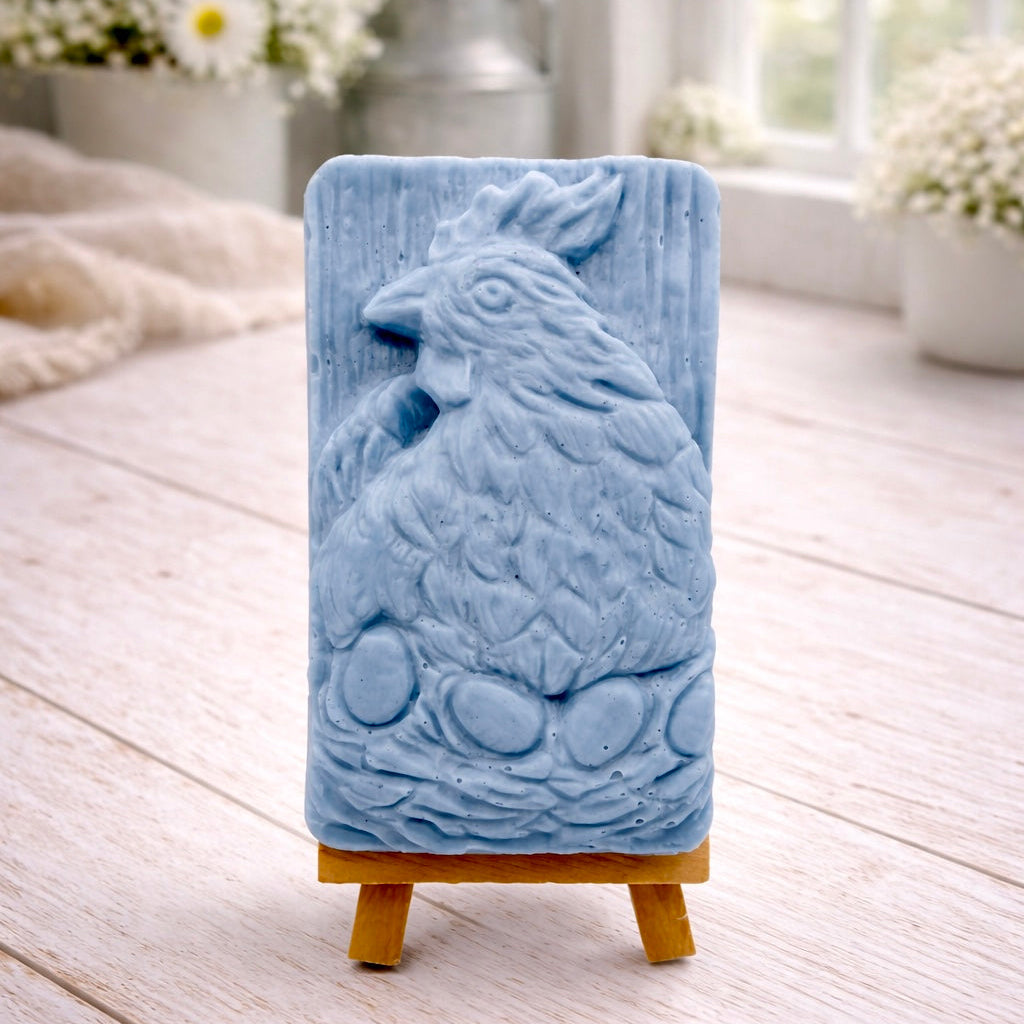 A rectangular handmade vegan soap in a natural colour, featuring a highly detailed relief carving of a broody hen chicken sitting on a nest of eggs, displayed upright on a light wooden surface.