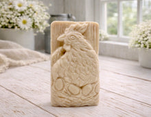 Load image into Gallery viewer, A rectangular handmade vegan soap in a natural colour, featuring a highly detailed relief carving of a broody hen chicken sitting on a nest of eggs, displayed upright on a light wooden surface.
