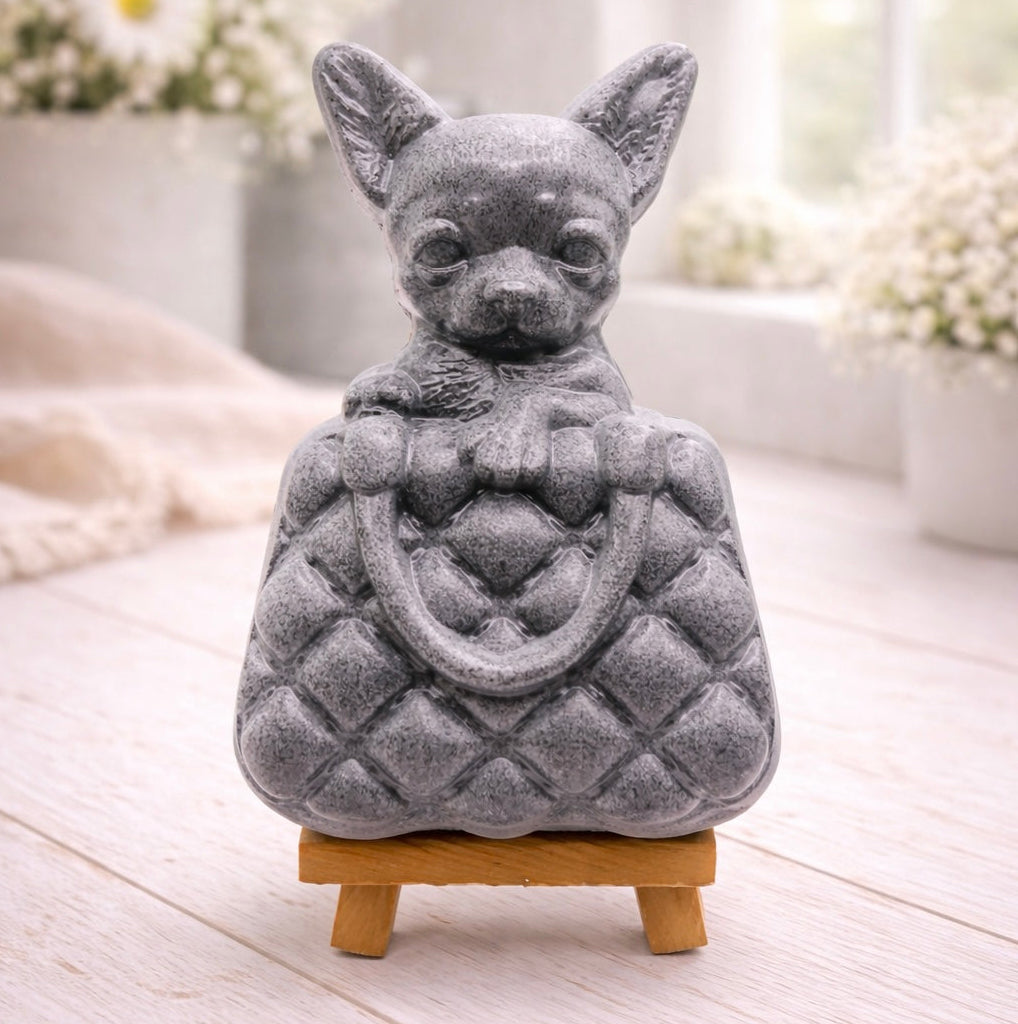 A highly detailed handmade vegan soap in a speckled grey marble finish, sculpted as a small chihuahua puppy with large ears peeking out from inside a quilted designer-style handbag, displayed on a miniature wooden easel.