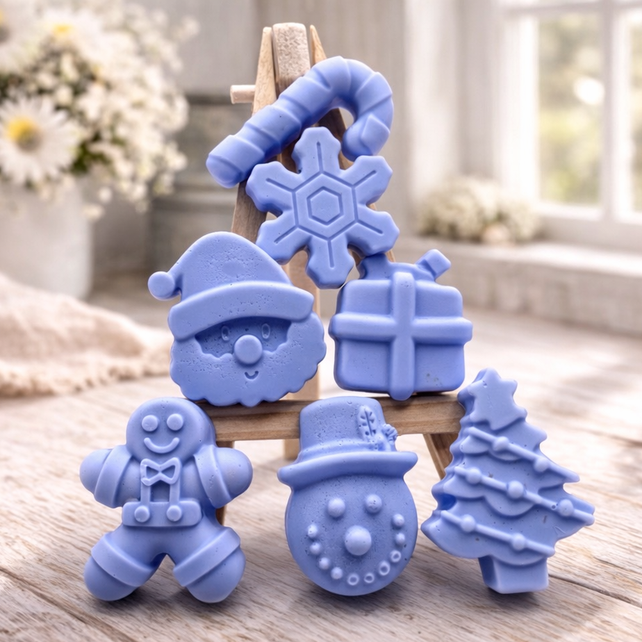 A collection of six handmade vegan Christmas soaps featuring a candy cane, snowflake, Santa Claus face, wrapped gift box, gingerbread man, snowman, and a decorated Christmas tree, arranged in a pyramid on a wooden stand.