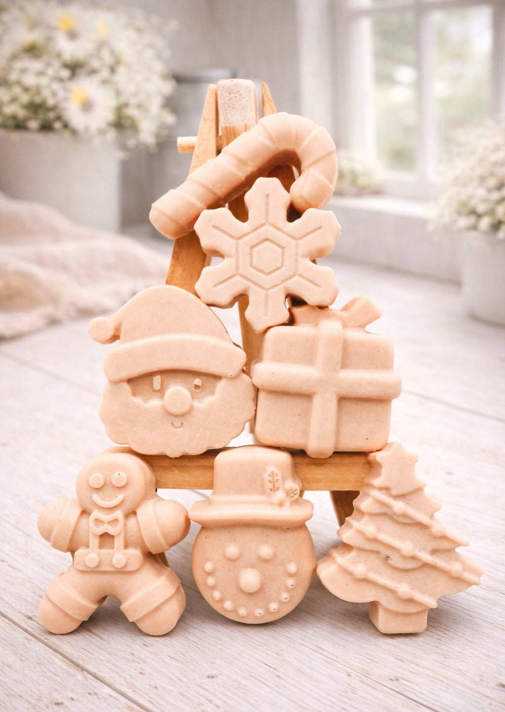 A collection of six handmade vegan Christmas soaps featuring a candy cane, snowflake, Santa Claus face, wrapped gift box, gingerbread man, snowman, and a decorated Christmas tree, arranged in a pyramid on a wooden stand.