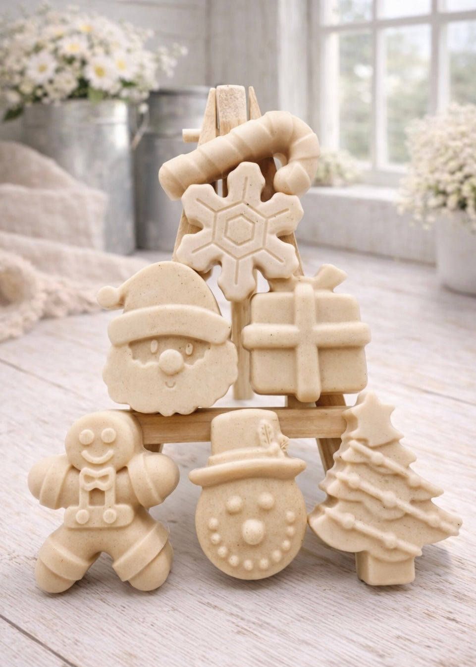 A collection of six handmade vegan Christmas soaps featuring a candy cane, snowflake, Santa Claus face, wrapped gift box, gingerbread man, snowman, and a decorated Christmas tree, arranged in a pyramid on a wooden stand.