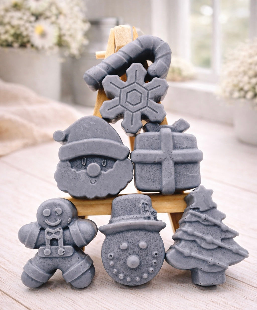 A collection of six handmade vegan Christmas soaps featuring a candy cane, snowflake, Santa Claus face, wrapped gift box, gingerbread man, snowman, and a decorated Christmas tree, arranged in a pyramid on a wooden stand.